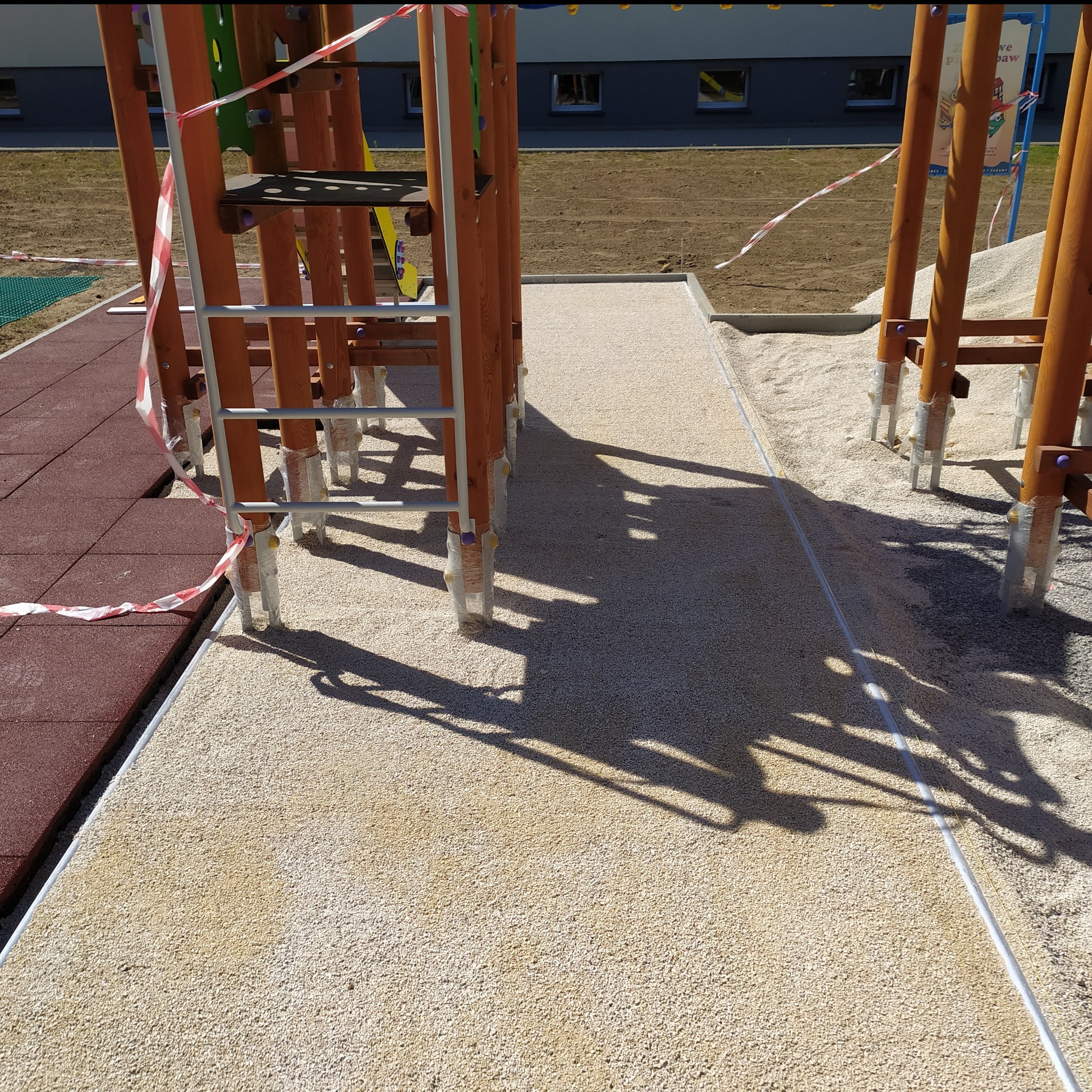 Nowa, jasna nawierzchnia z drobnego kruszywa na placu zabaw, z elementami drewnianej konstrukcji zabezpieczonymi folią i taśmą ostrzegawczą.