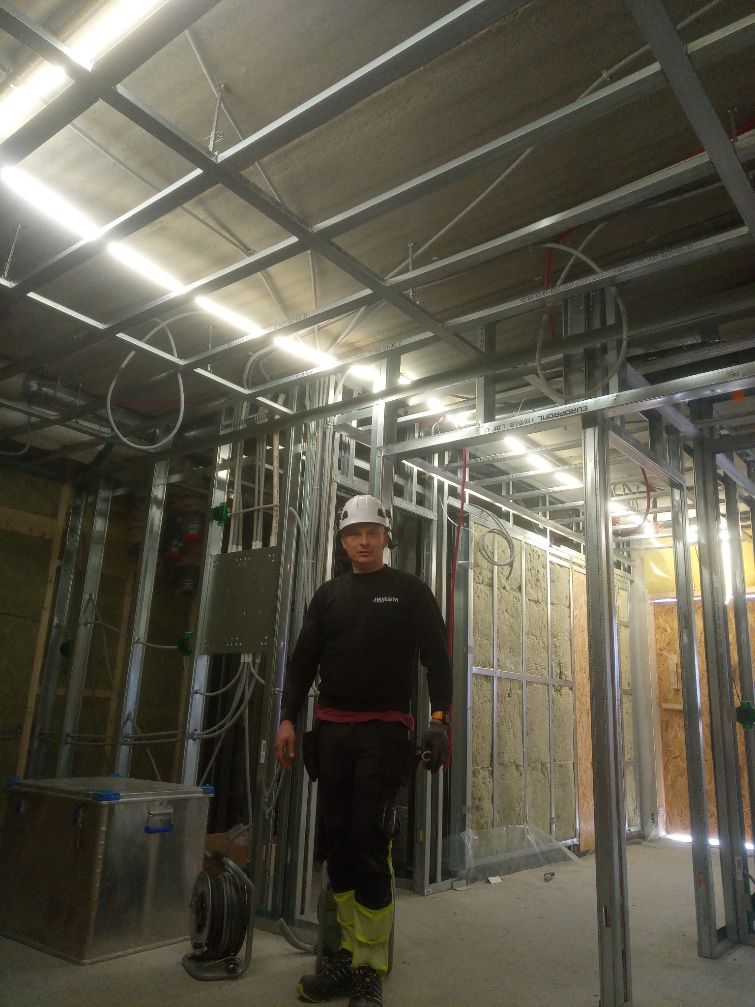Pracownik w kasku i roboczym ubraniu stoi wewnątrz pomieszczenia w trakcie budowy, widoczne metalowe profile konstrukcyjne ścian i sufitu, instalacje elektryczne i izolacja z wełny mineralnej.