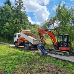 AP-KOP - Pomarańczowa minikoparka Kubota z niebieskim osprzętem kopiąca rów obok białej wywrotki na tle zieleni