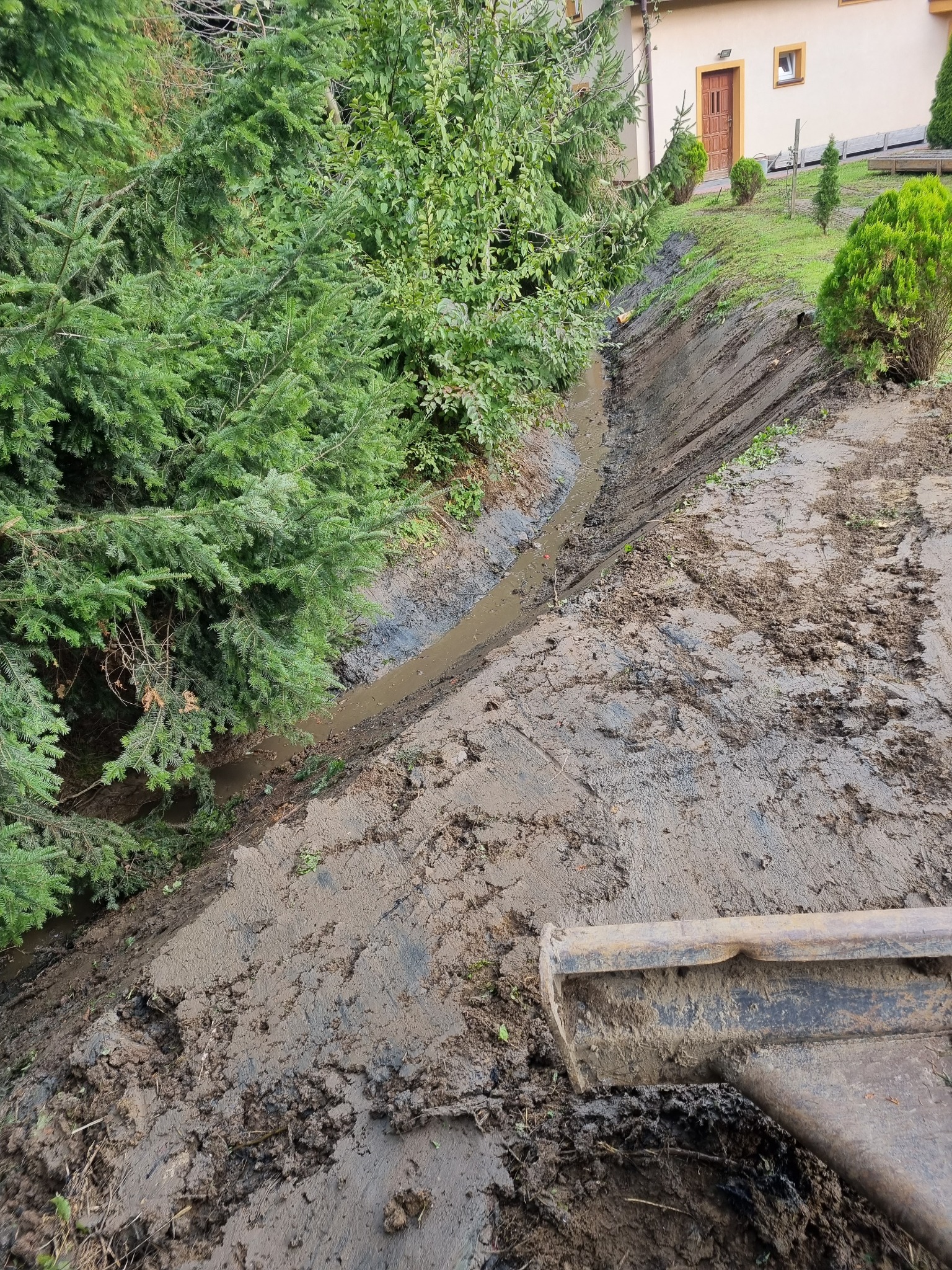 Wykopany rów wypełniony wodą, ziemia po bokach, widoczna łyżka koparki w prawym dolnym rogu, w tle drzewa i budynek.