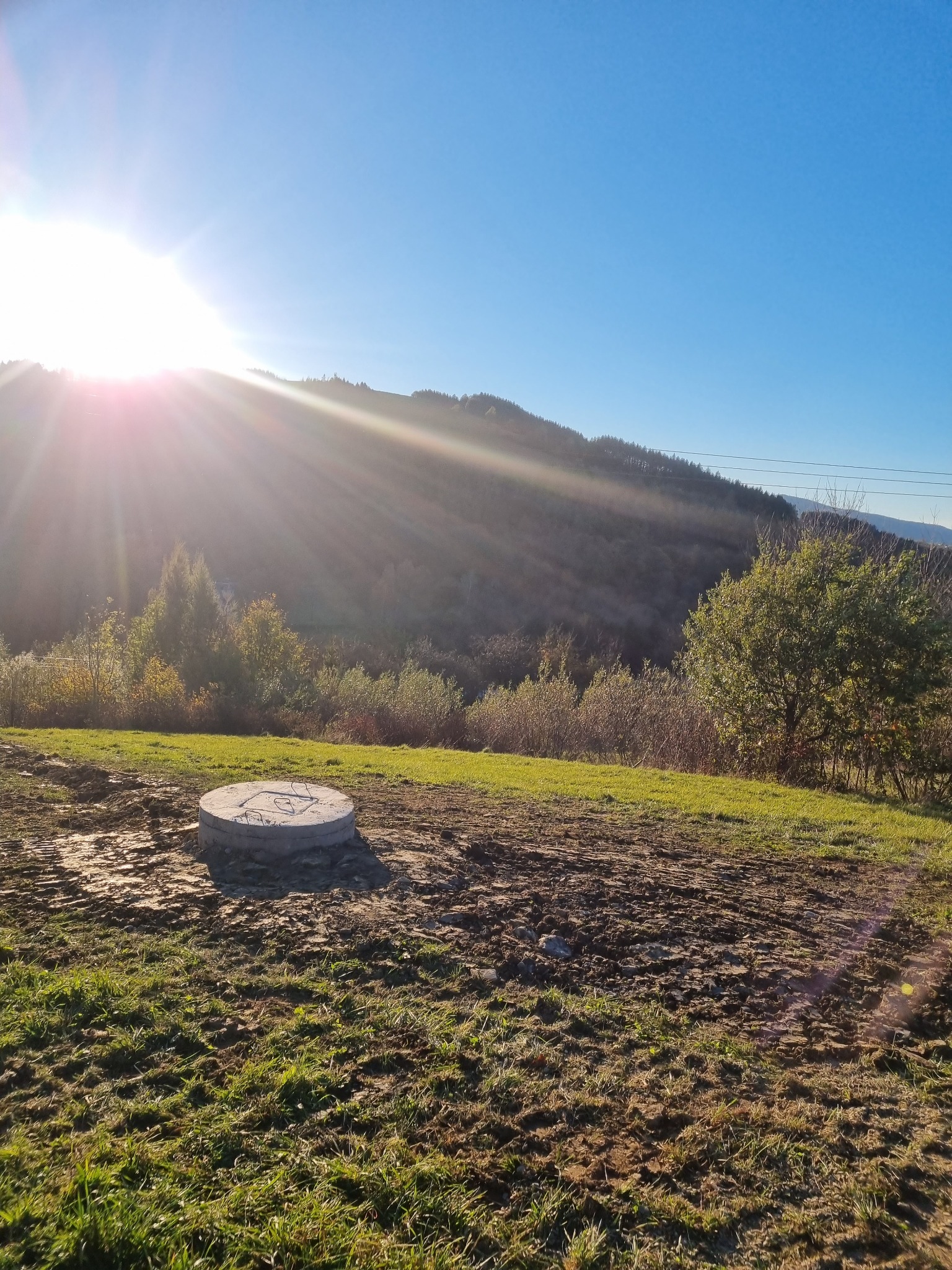 Betonowa pokrywa szamba na nierównym, częściowo wykopanym terenie z widokiem na zalesione wzgórze pod jasnym słońcem.