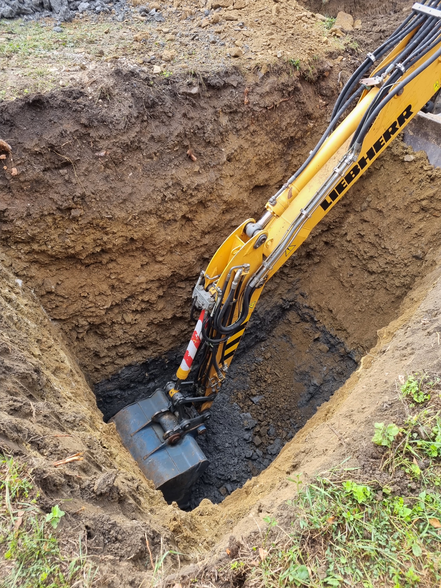 Żółta koparka Liebherr pogłębia kwadratowy wykop w ziemi, widoczne warstwy gleby o różnej barwie.