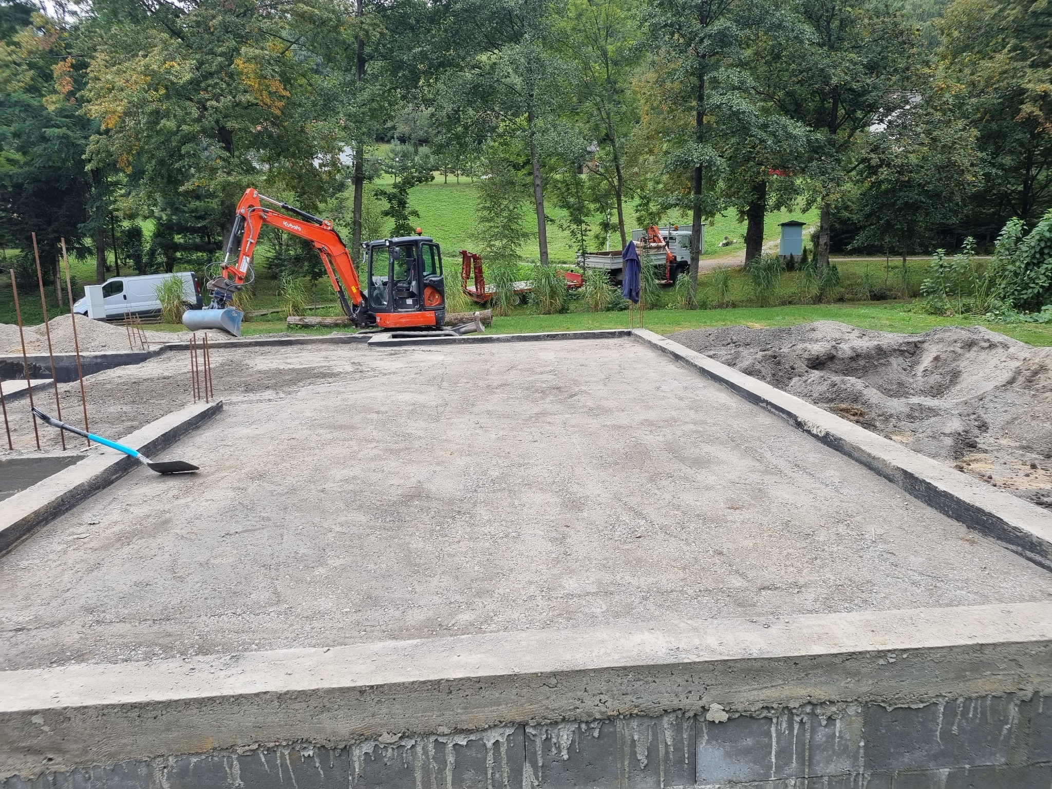 Wylany fundament pod budynek, w tle pomarańczowa koparka Kubota i inne maszyny budowlane oraz biały bus na tle zieleni.