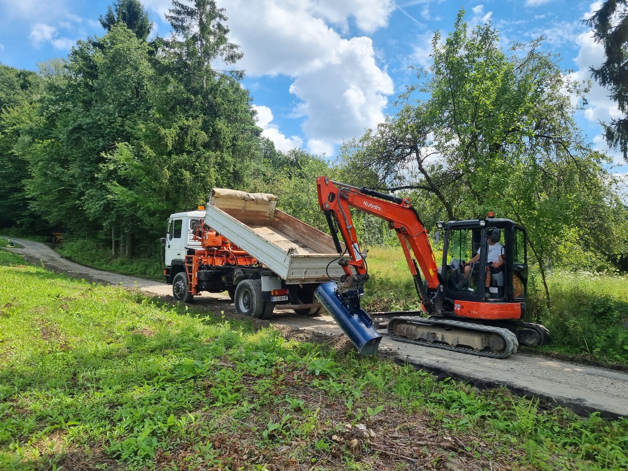 Pomarańczowa minikoparka Kubota z niebieskim osprzętem kopiąca rów obok białej wywrotki na tle zieleni