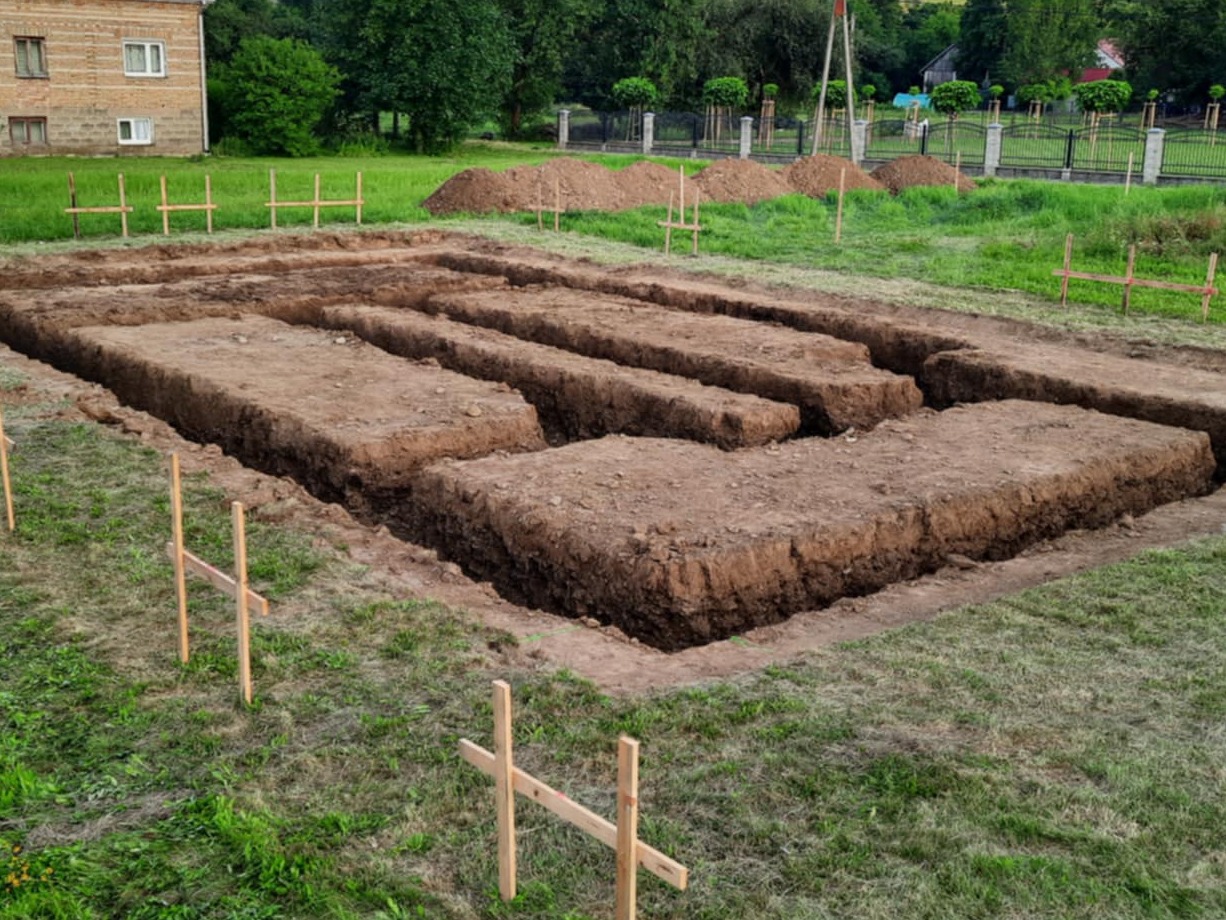 Przygotowany wykop pod fundamenty w Nowym Żmigrodzie. Widoczne równe rowy w ziemi, drewniane znaczniki i pryzmy ziemi w tle. Prace ziemne.