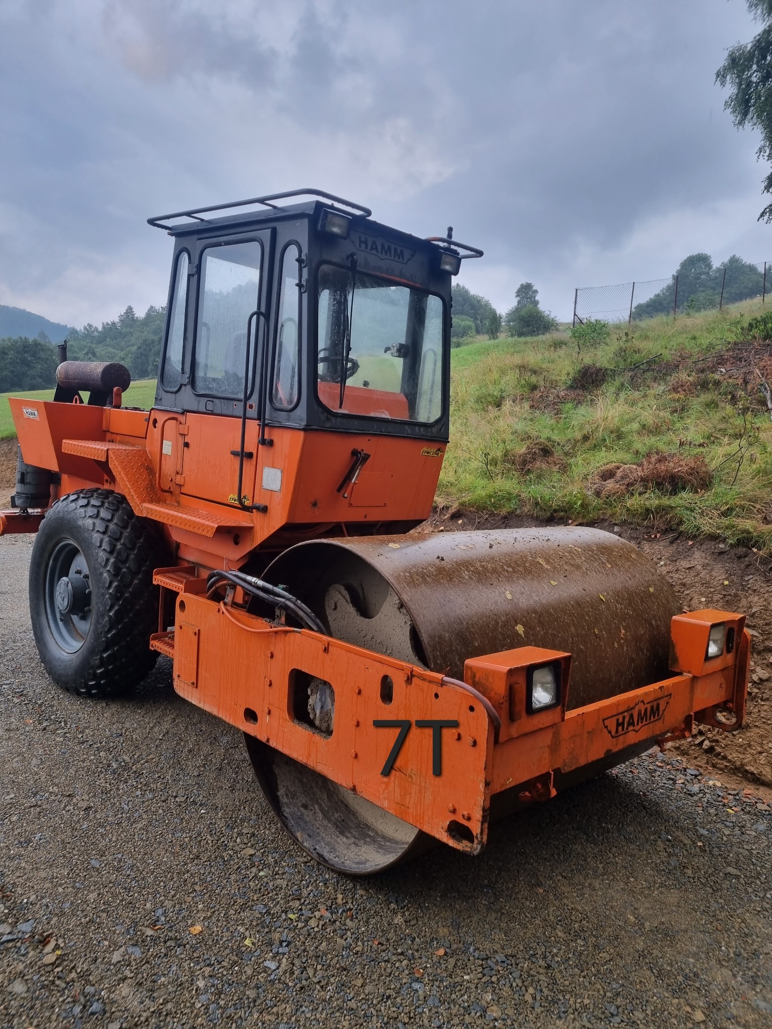 Pomarańczowy walec drogowy Hamm na żwirowej drodze, w tle zielone wzgórza pod pochmurnym niebem. Maszyna z kabiną, widoczne detale i logo producenta.