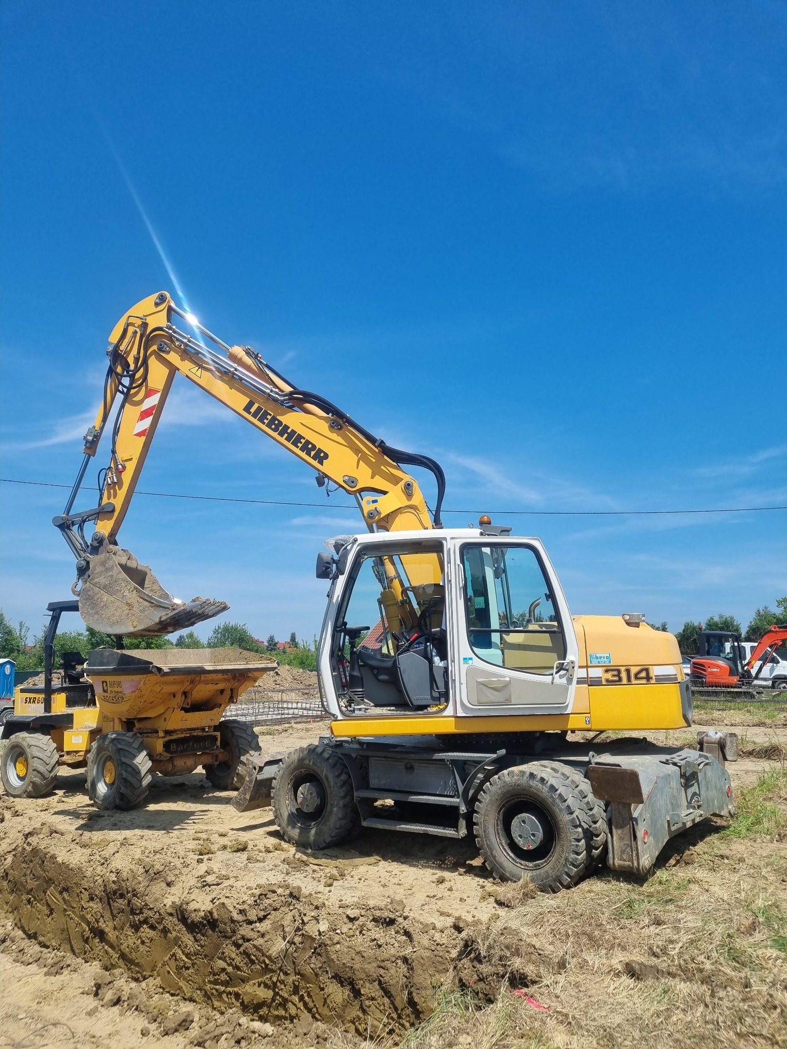 Żółta koparka Liebherr 314 nabiera ziemię do wywrotki Barford na tle wykopu i błękitnego nieba. Prace ziemne w toku, widoczne inne maszyny budowlane w tle.