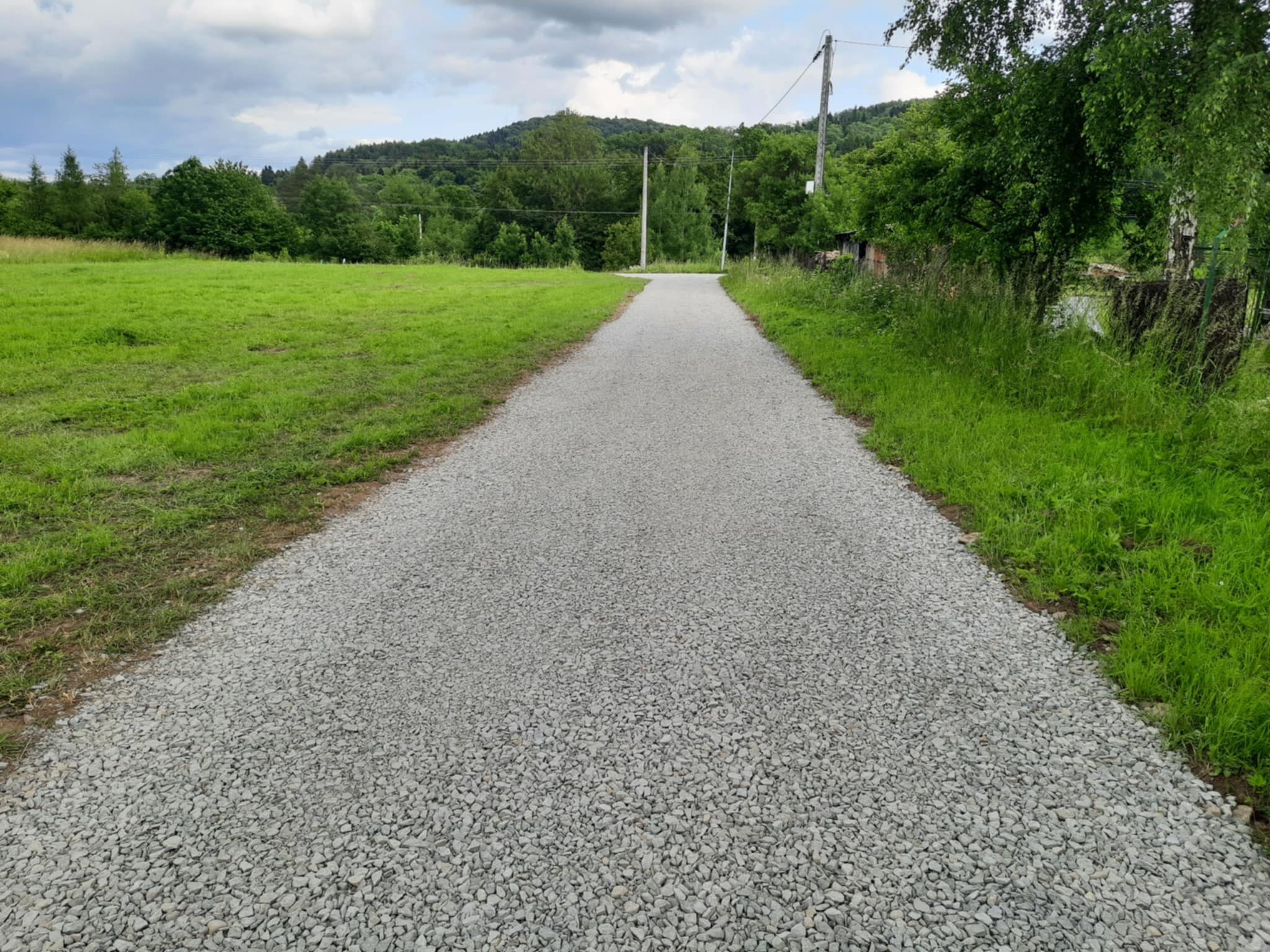 Utwardzona, szara, żwirowa droga polna prowadząca przez zielony teren w kierunku zalesionego wzgórza pod pochmurnym niebem. Widoczne słupy energetyczne.