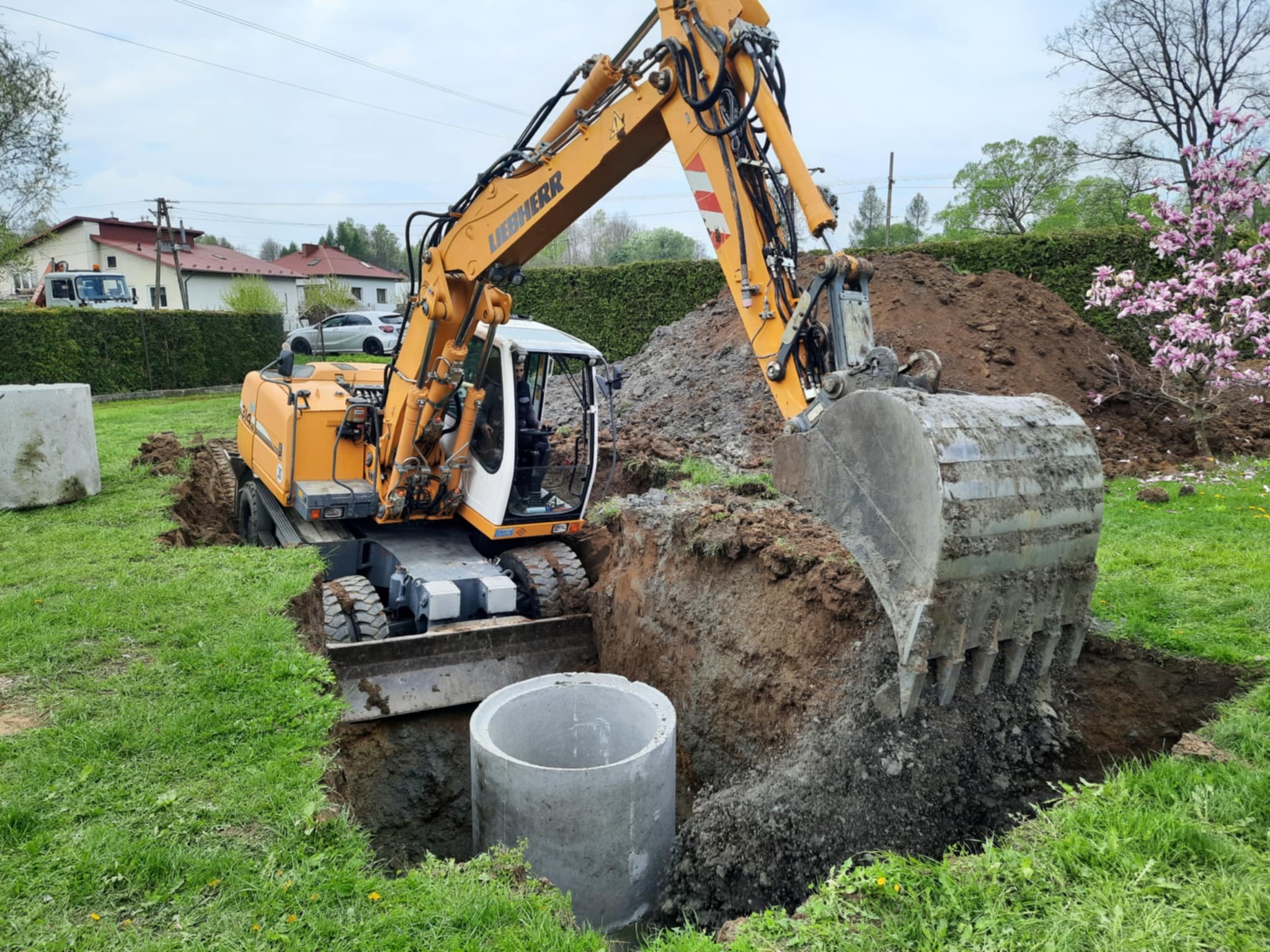 Żółta koparka Liebherr w trakcie wykopu pod betonowy krąg studzienny na zielonym trawniku, z widocznym operatorem i stertą ziemi w tle.