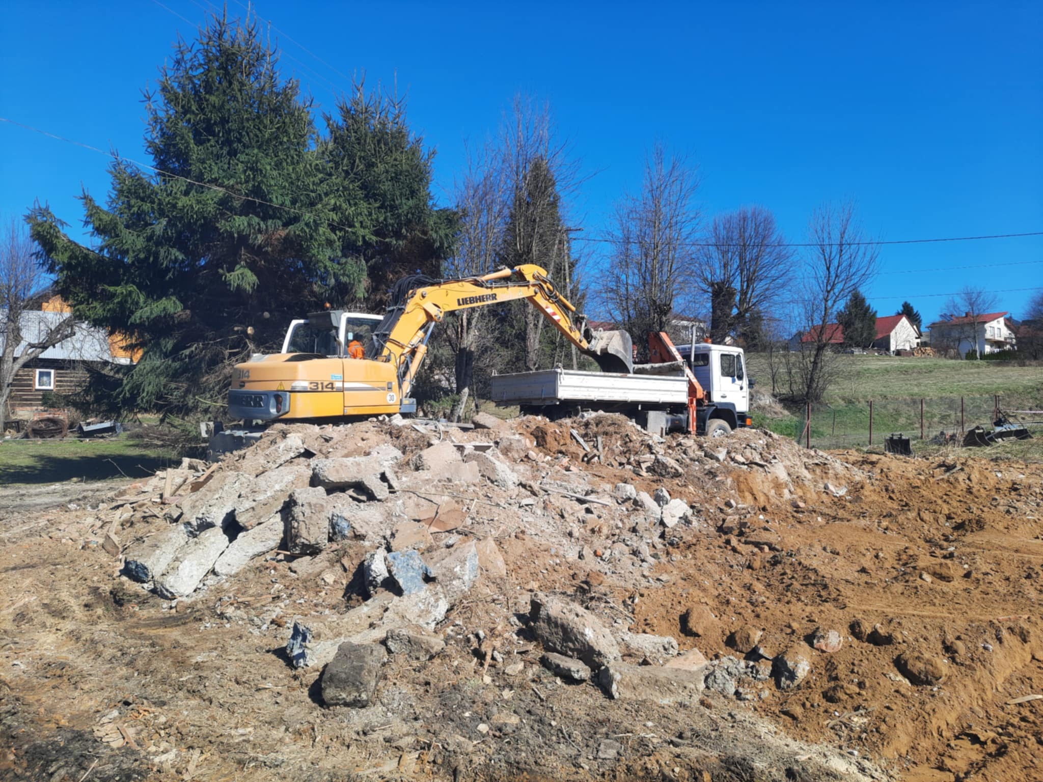 Żółta koparka Liebherr ładuje gruz do białej ciężarówki na tle błękitnego nieba i zabudowań. Widoczne fragmenty zniszczonych elementów betonowych.