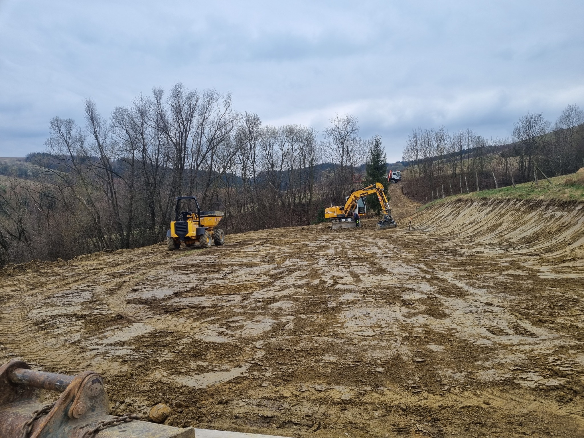 Żółta koparka i wywrotka pracują na nierównym, błotnistym terenie. W tle drzewa bez liści i pagórkowaty krajobraz pod pochmurnym niebem. Widoczne ślady opon.
