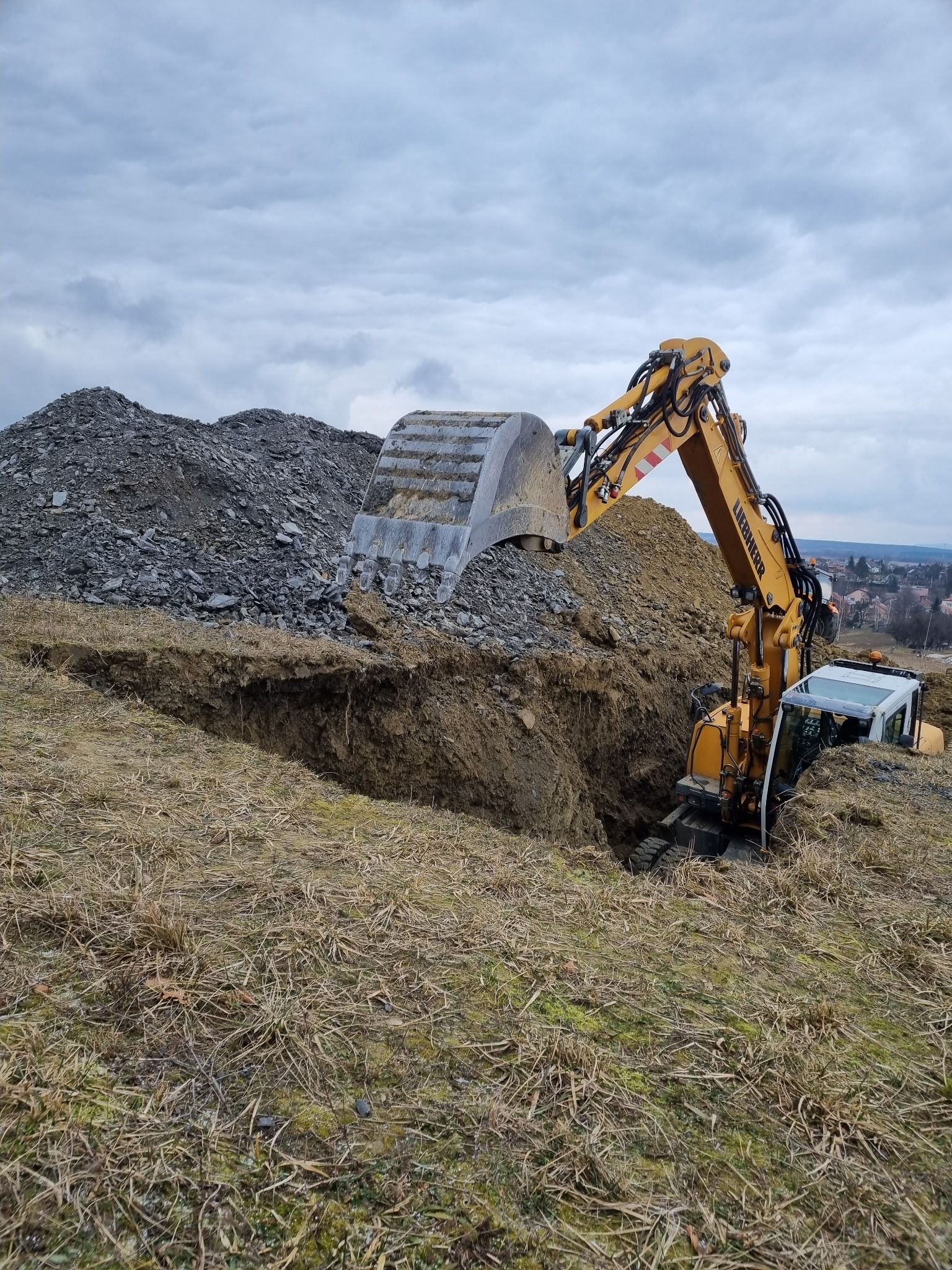 Żółta koparka Liebherr w trakcie wykopu ziemi, z hałdą gruzu w tle i widokiem na zabudowania w oddali, pochmurne niebo.