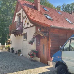 Dwukondygnacyjny dom z czerwonym dachem, drewnianą elewacją i dekoracyjnymi elementami ludowymi, widoczny z poziomu brukowanej ulicy, z zaparkowanym obok samochodem dostawczym.
