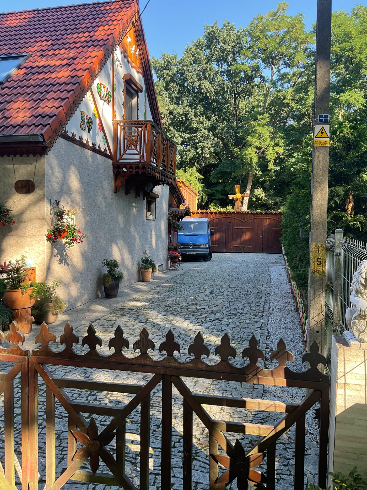 Ujęcie domu z czerwonym dachem, zdobieniami na ścianie, drewnianym balkonem, kwiatami w donicach, brukowanym podjazdem i niebieskim samochodem dostawczym zaparkowanym przed drewnianą bramą...