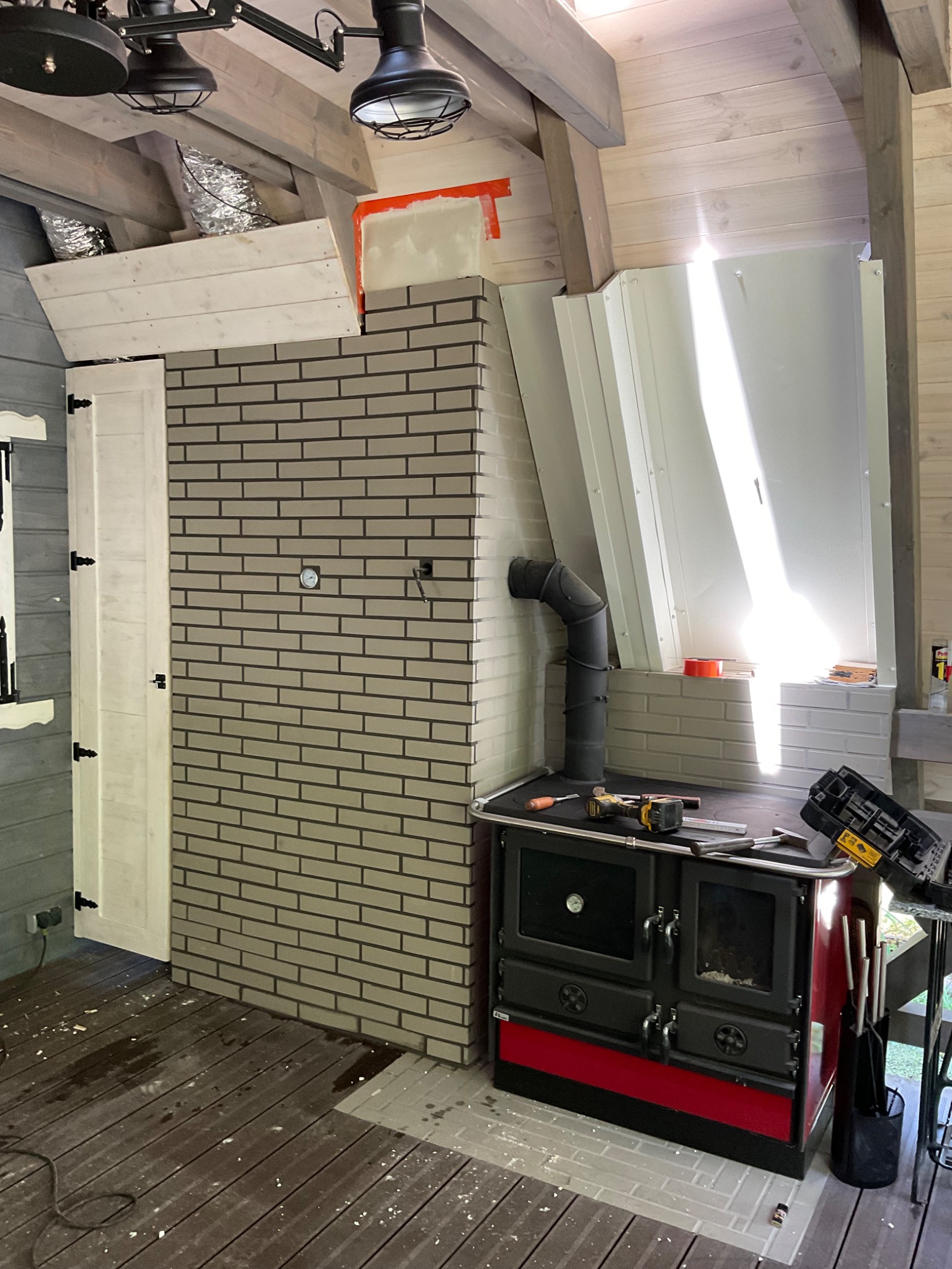 Wnętrze z kominkiem kaflowym w trakcie montażu, widoczne narzędzia, fragmenty obudowy i elementy instalacji kominowej, surowy stan wykończenia poddasza.