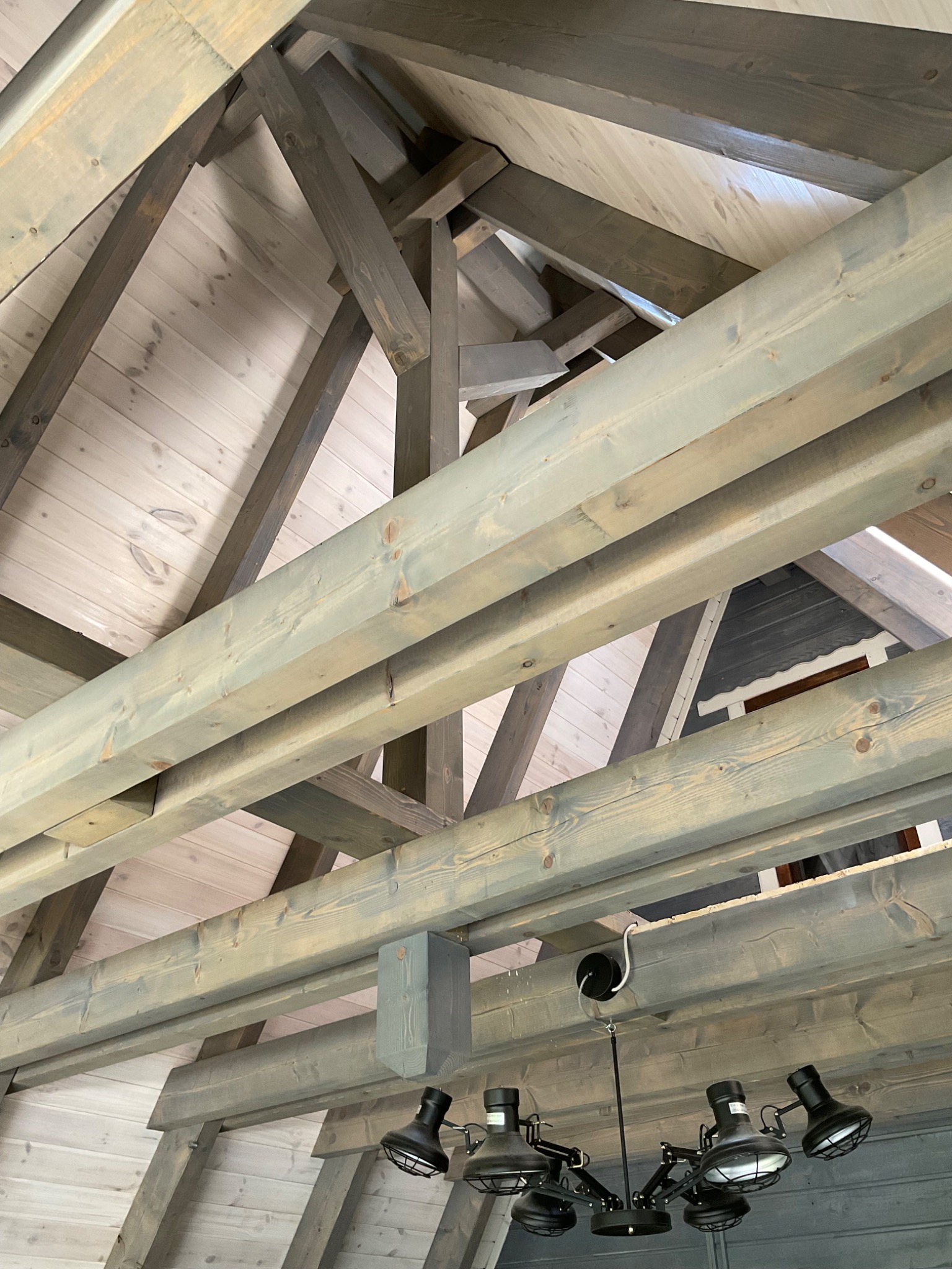 Drewniana konstrukcja dachu poddasza z widocznymi belkami nośnymi w odcieniach szarości i bieli, oświetlona lampą w stylu industrialnym.