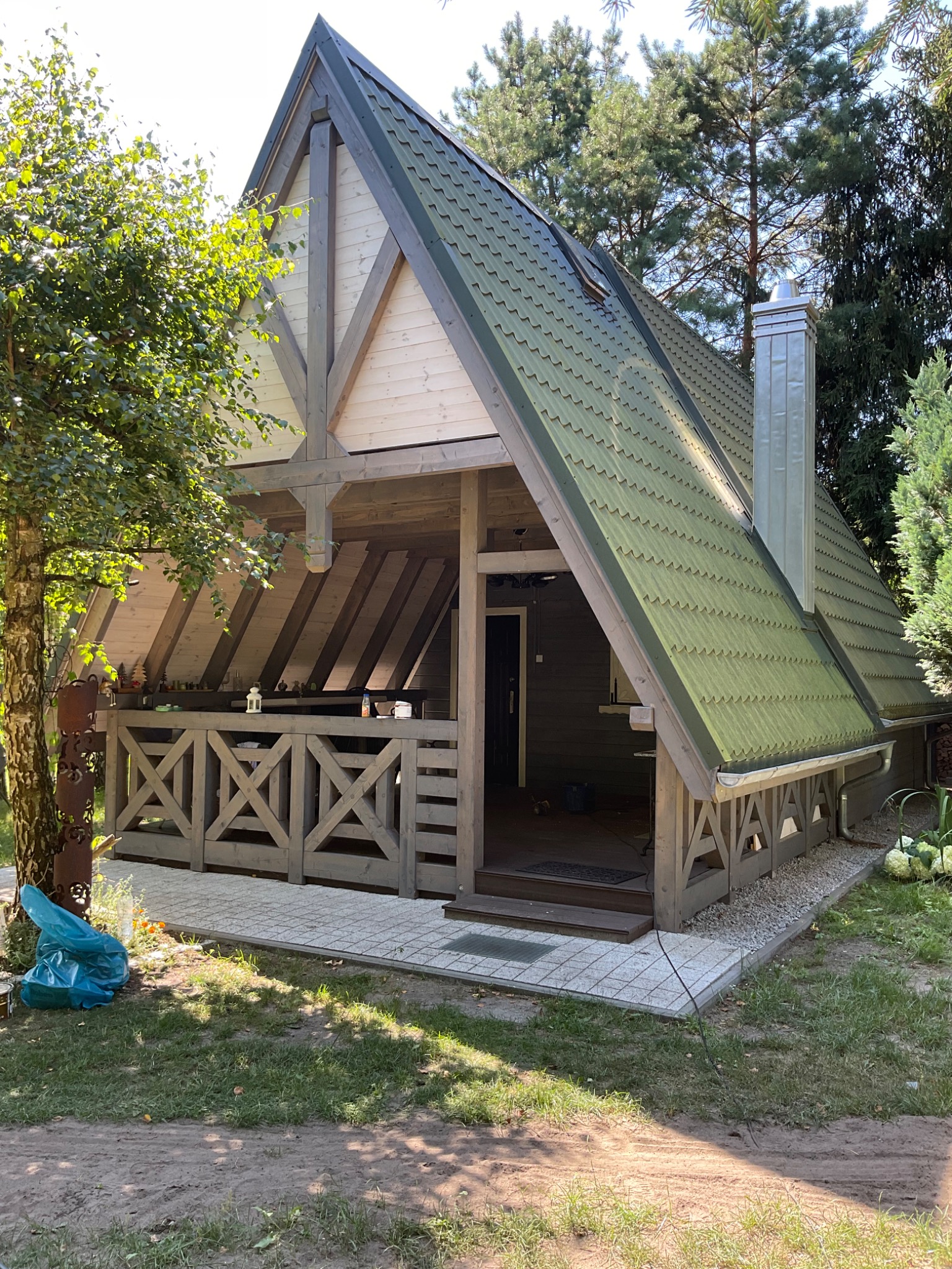 Drewniana altana z zielonym dachem blaszanym, wykończona w stylu skandynawskim, z ozdobną balustradą i kominem ze stali nierdzewnej, otoczona zielenią.