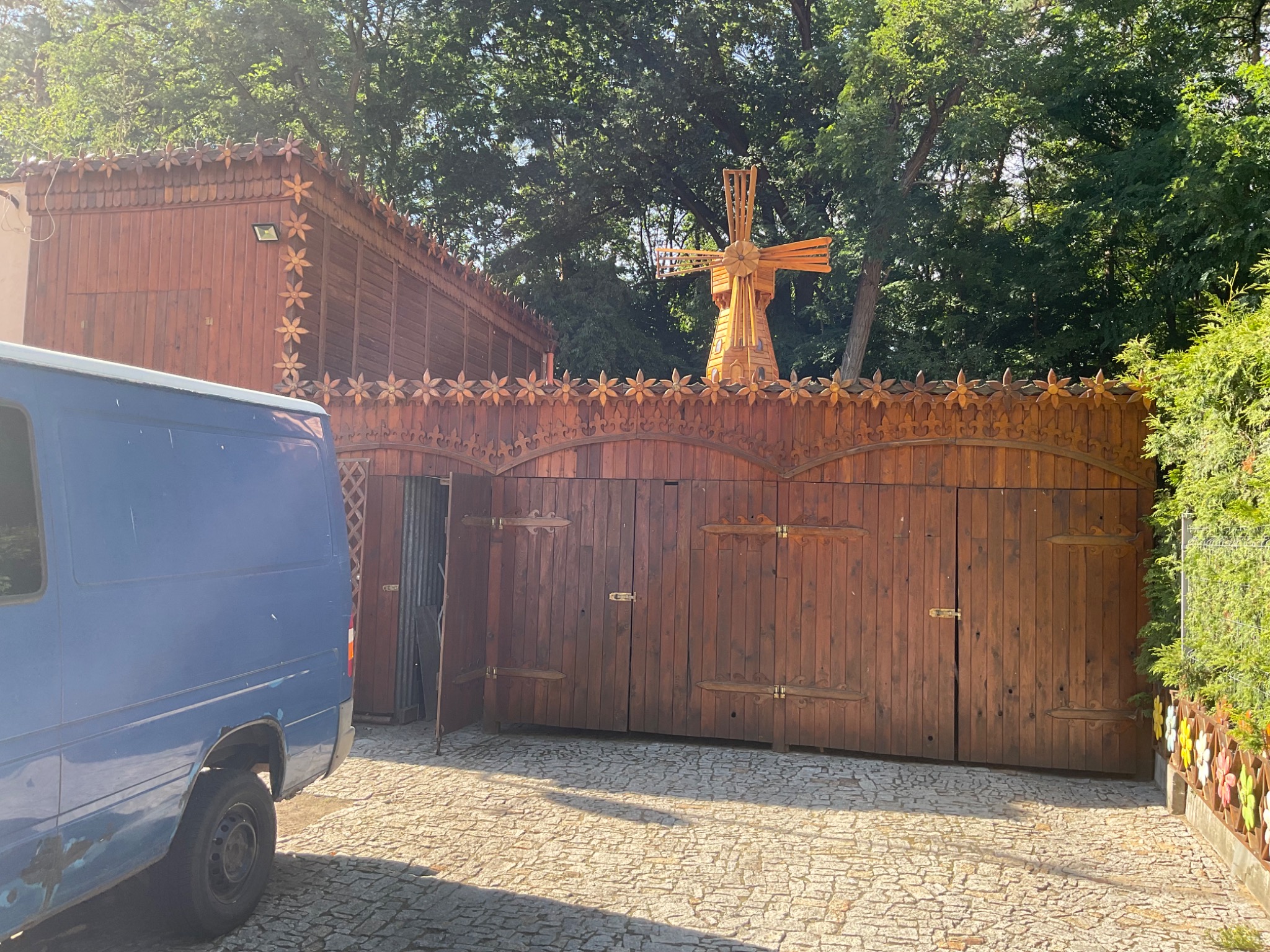 Drewniana brama z dekoracyjnymi kwiatami i wiatrakiem na tle zieleni, obok zaparkowany niebieski bus.