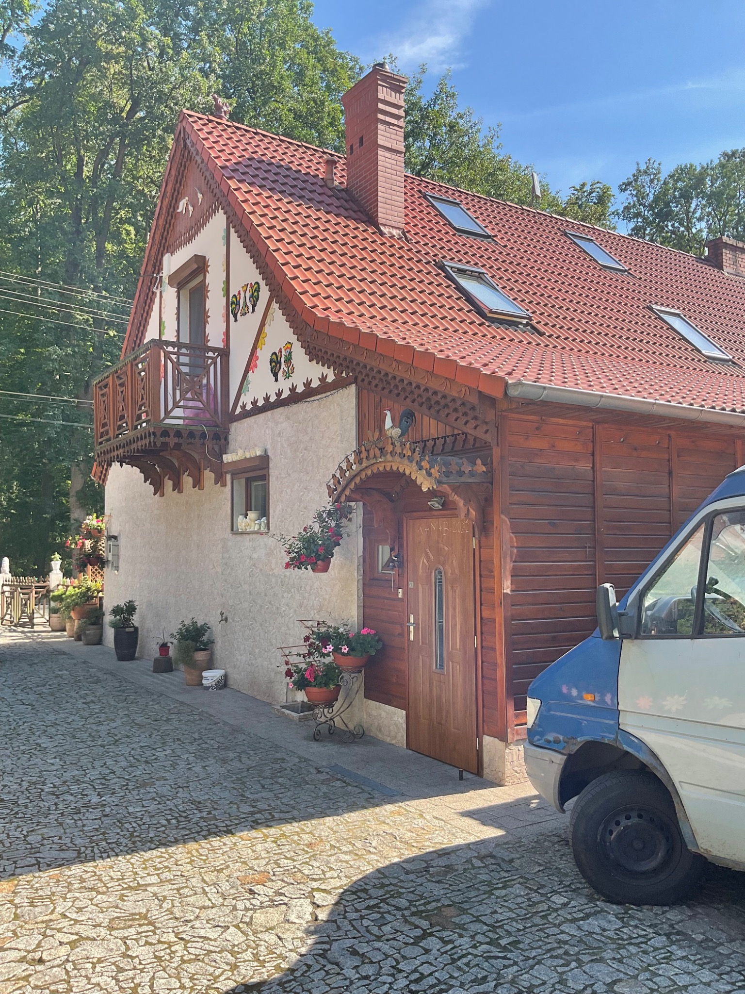 Dwukondygnacyjny dom z czerwonym dachem, drewnianą elewacją i dekoracyjnymi elementami ludowymi, widoczny z poziomu brukowanej ulicy, z zaparkowanym obok samochodem dostawczym.