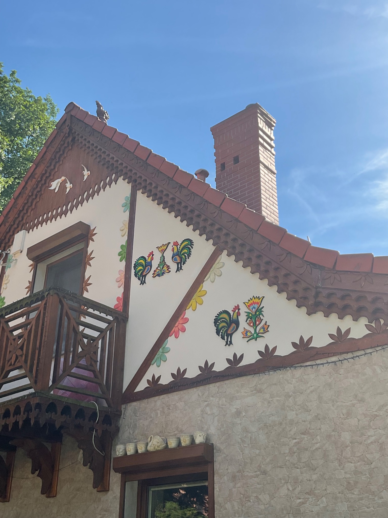 Fasada domu z dekoracyjnymi elementami: rzeźbione drewniane wykończenia pod dachem, malowane koguty i kwiaty, balkon z drewnianą balustradą, oraz ceglany komin na tle błękitnego nieba.