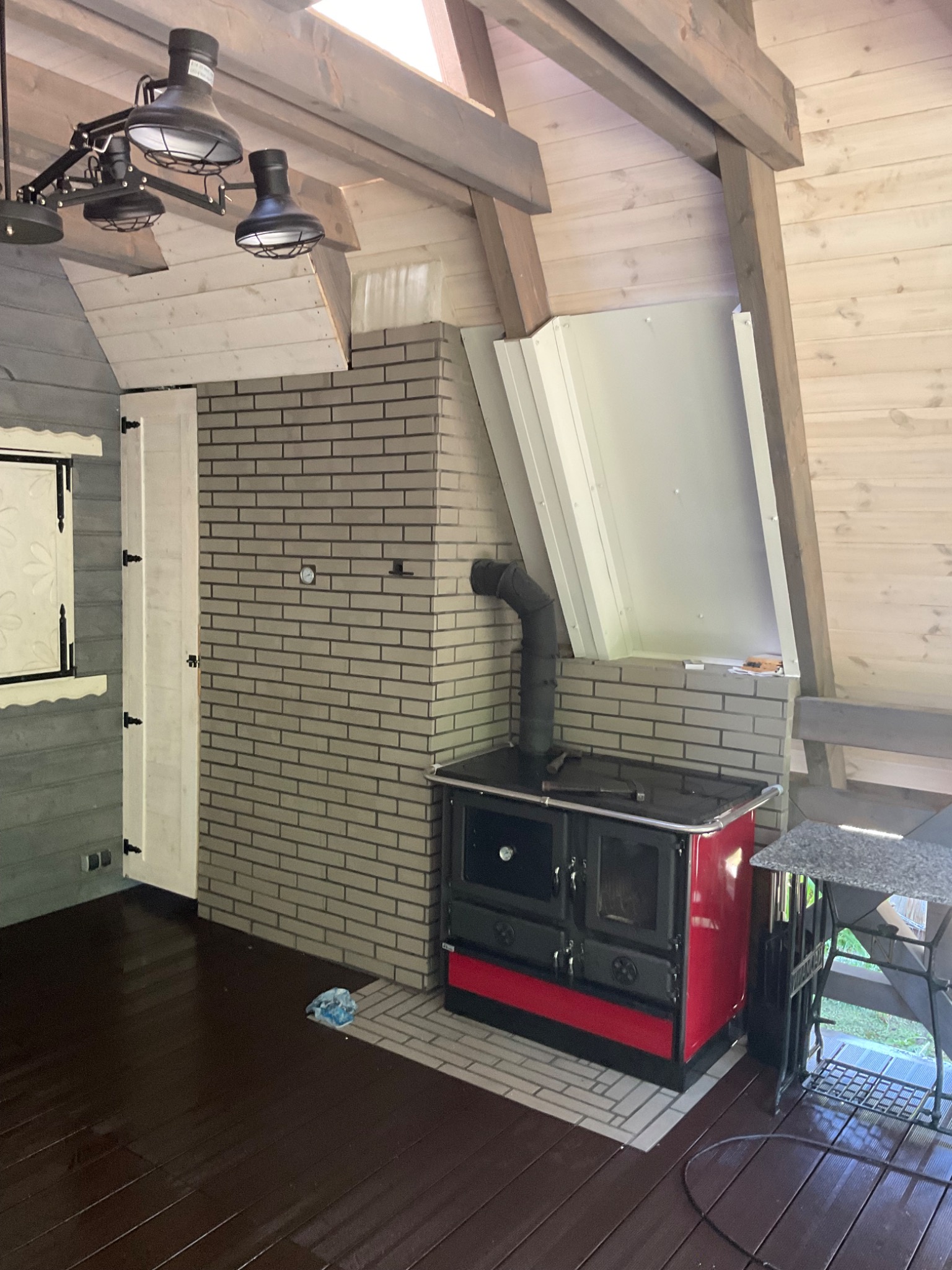 Wnętrze z drewnianą boazerią i skośnym sufitem, piec kaflowy z czerwonym wykończeniem, murowany komin z szarej cegły, industrialne lampy sufitowe i widoczny fragment okna.