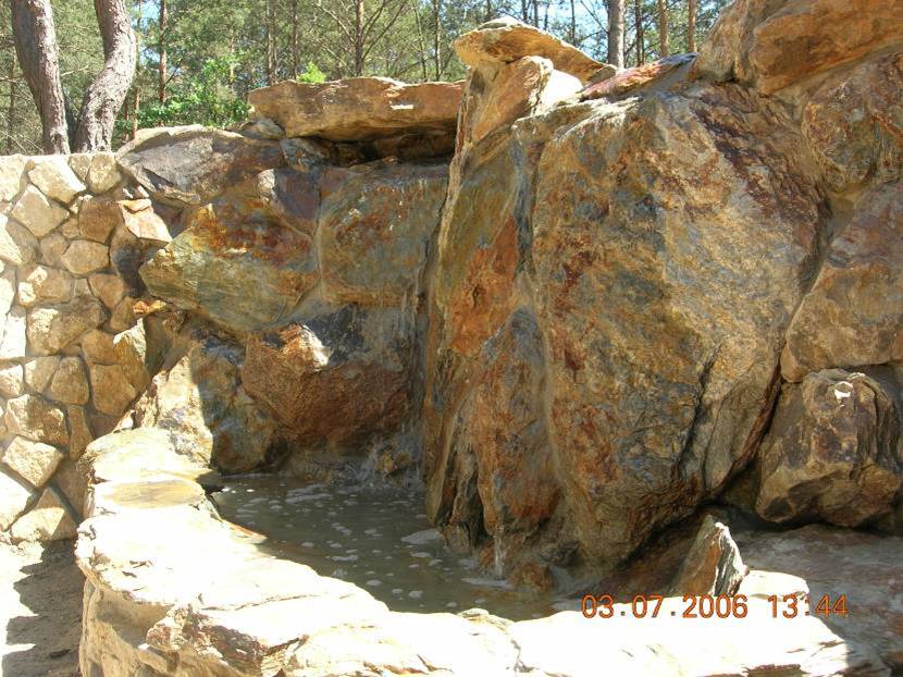 Kamienna konstrukcja z wodą, imitująca naturalny wodospad w ogrodzie, wykonana z nieregularnych kamieni o różnej wielkości i odcieniach brązu i szarości, otoczona drzewami.