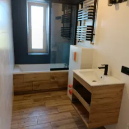 Nowoczesna łazienka z drewnopodobnymi płytkami podłogowymi, zabudową wanny z drewnianymi panelami, umywalką z szafką wiszącą w stylu drewna i czarną armaturą, oraz grzejnikiem drabinkowym.