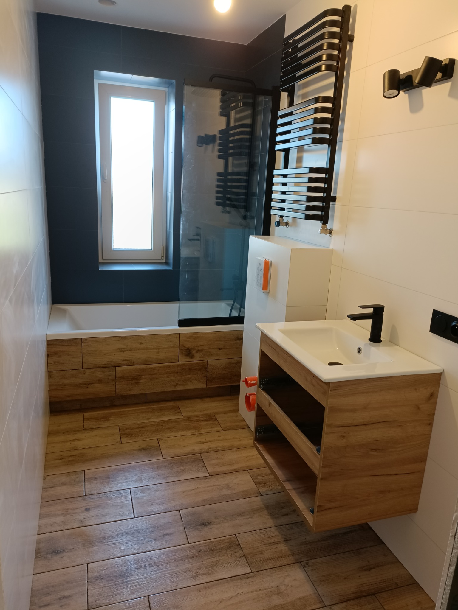 Nowoczesna łazienka z drewnopodobnymi płytkami podłogowymi, zabudową wanny z drewnianymi panelami, umywalką z szafką wiszącą w stylu drewna i czarną armaturą, oraz grzejnikiem drabinkowym.