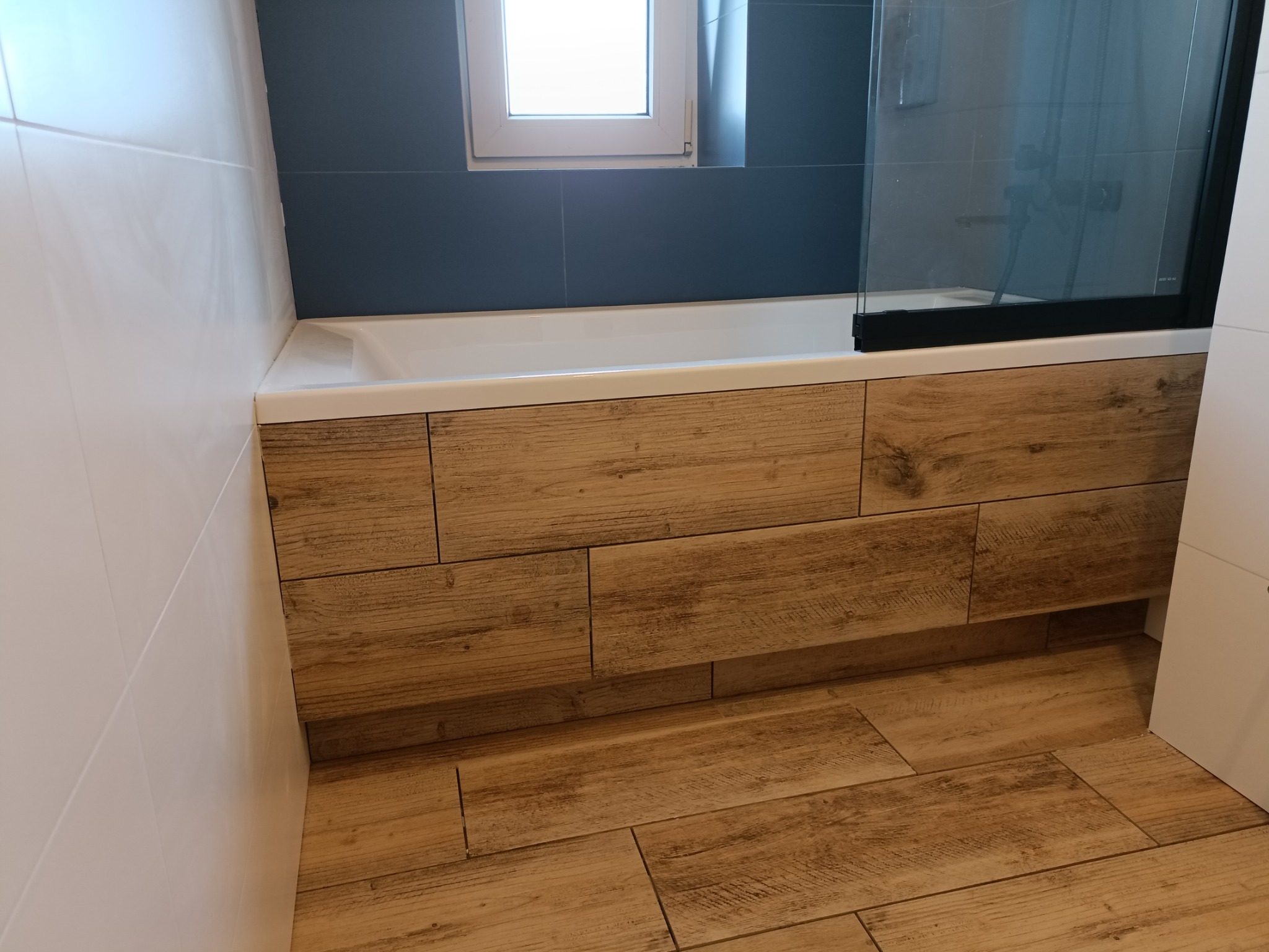 Wykończona łazienka z drewnopodobnymi płytkami na podłodze i obudowie wanny, białe płytki na ścianach oraz niebieska ściana z oknem, kabina prysznicowa z czarnym obramowaniem.