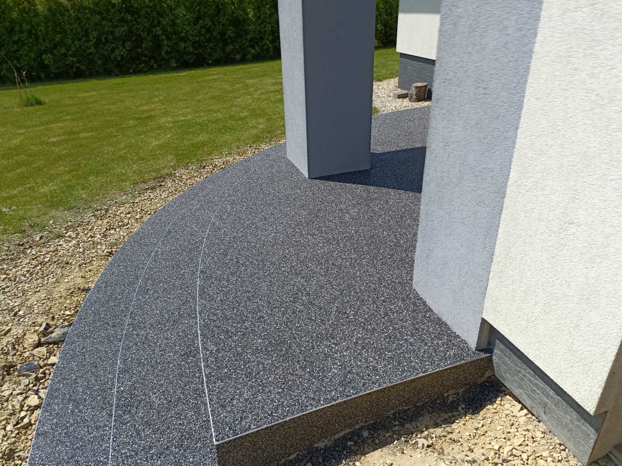Nowoczesna, żywiczna posadzka zewnętrzna z drobnym kruszywem w odcieniach szarości, przylegająca do elewacji budynku z kolumnami, otoczona żwirem i trawnikiem.