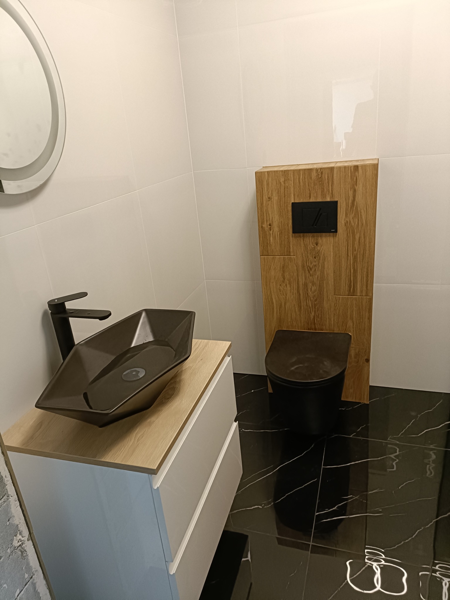 Nowoczesna łazienka z czarną armaturą: umywalka w kształcie trapezu, bateria, wisząca miska WC z drewnianą obudową, białe płytki ścienne i czarna marmurowa podłoga.