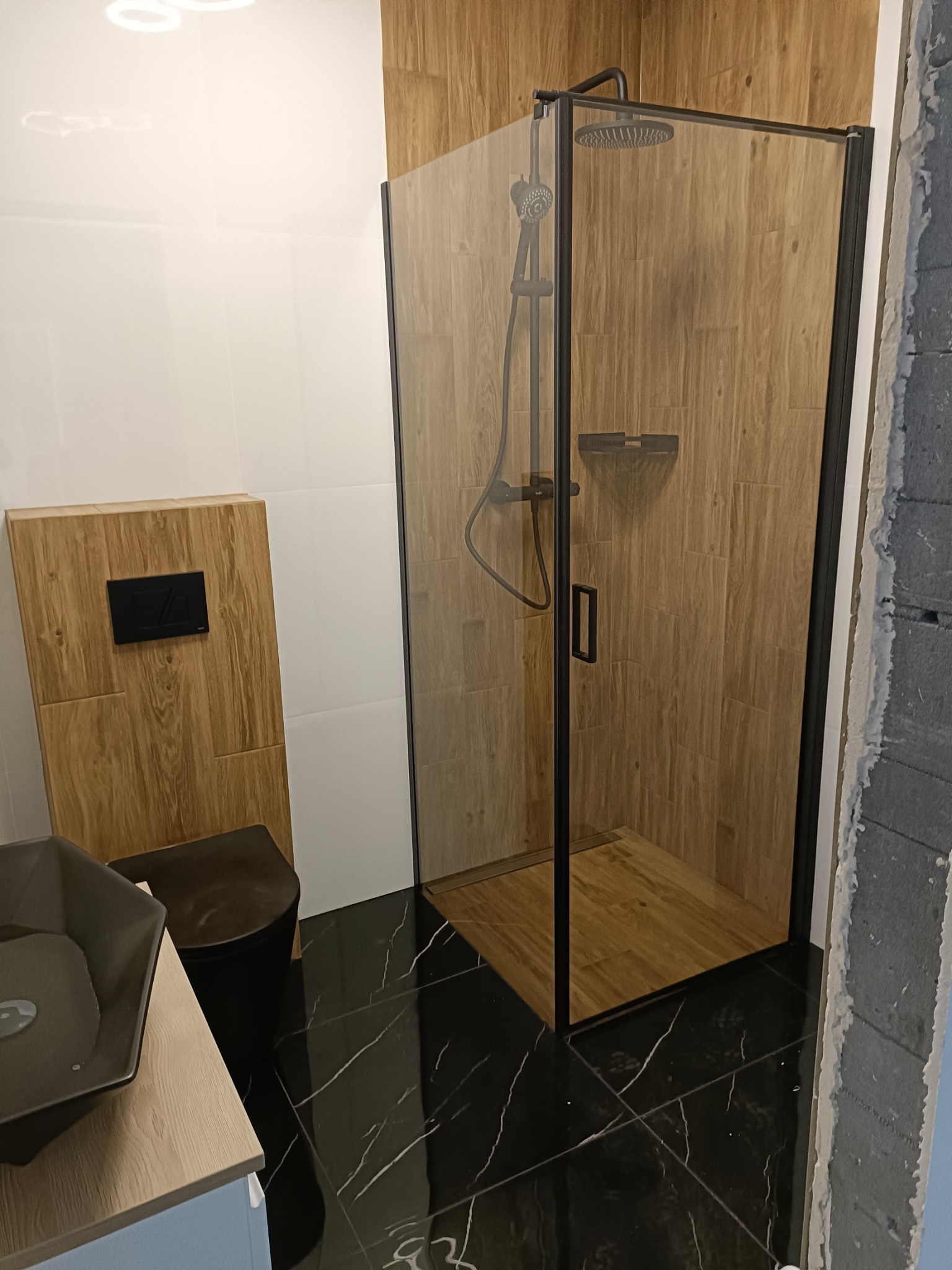 Nowoczesna łazienka z czarną armaturą, kabiną prysznicową typu walk-in z drewnopodobnymi płytkami, czarną ceramiką i błyszczącą czarną podłogą imitującą marmur.