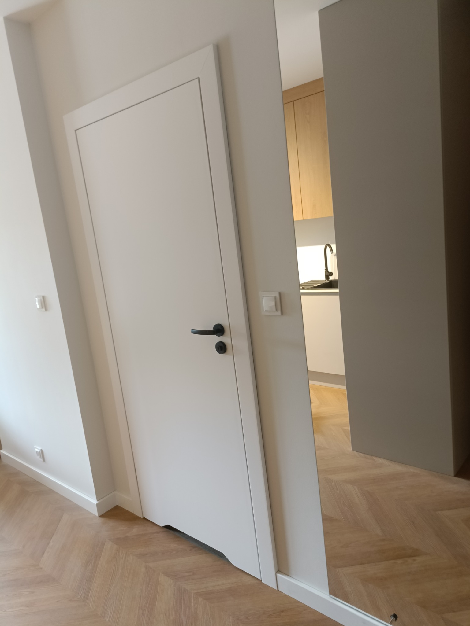 Białe drzwi wewnętrzne z czarną klamką, widok na fragment kuchni z drewnianymi szafkami i czarnym zlewem, parkiet ułożony w jodełkę.