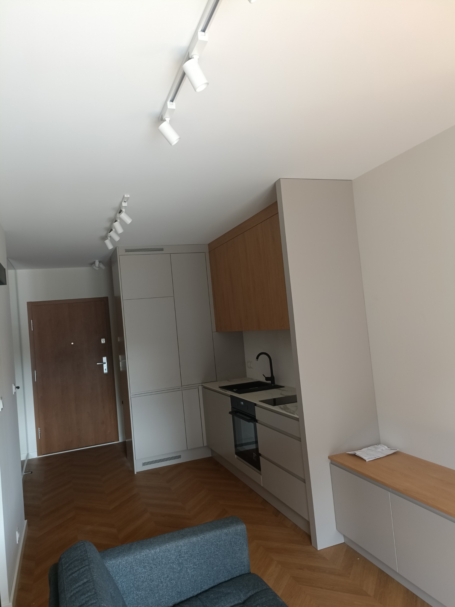 Nowoczesne wnętrze mieszkania z aneksem kuchennym, drewnianą podłogą ułożoną w jodełkę i oświetleniem punktowym na szynoprzewodach.