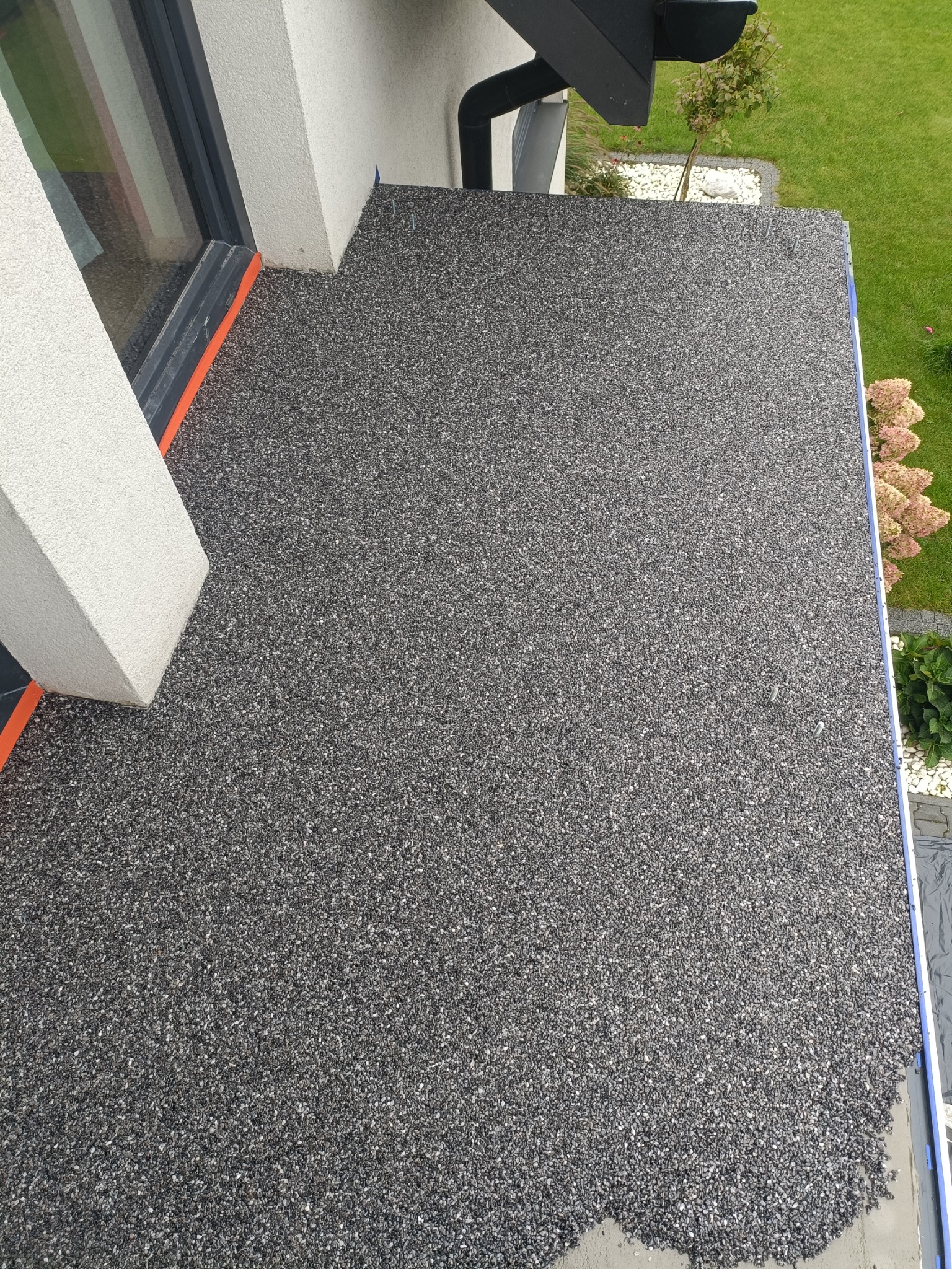 Wykończony taras z żywicy epoksydowej z widocznymi detalami, takimi jak listwy wykończeniowe, fragment okna i elewacji budynku oraz elementy ogrodu w tle.