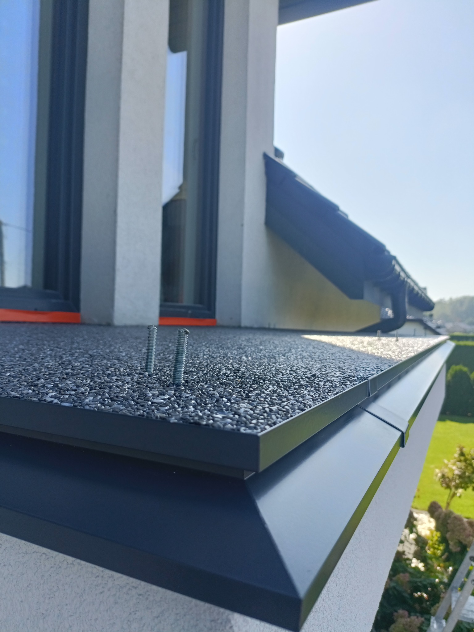 Wykończony fragment dachu z papy z posypką, widoczne dwa wystające wkręty, okno z pomarańczową uszczelką, rynna i fragment elewacji budynku w słoneczny dzień.