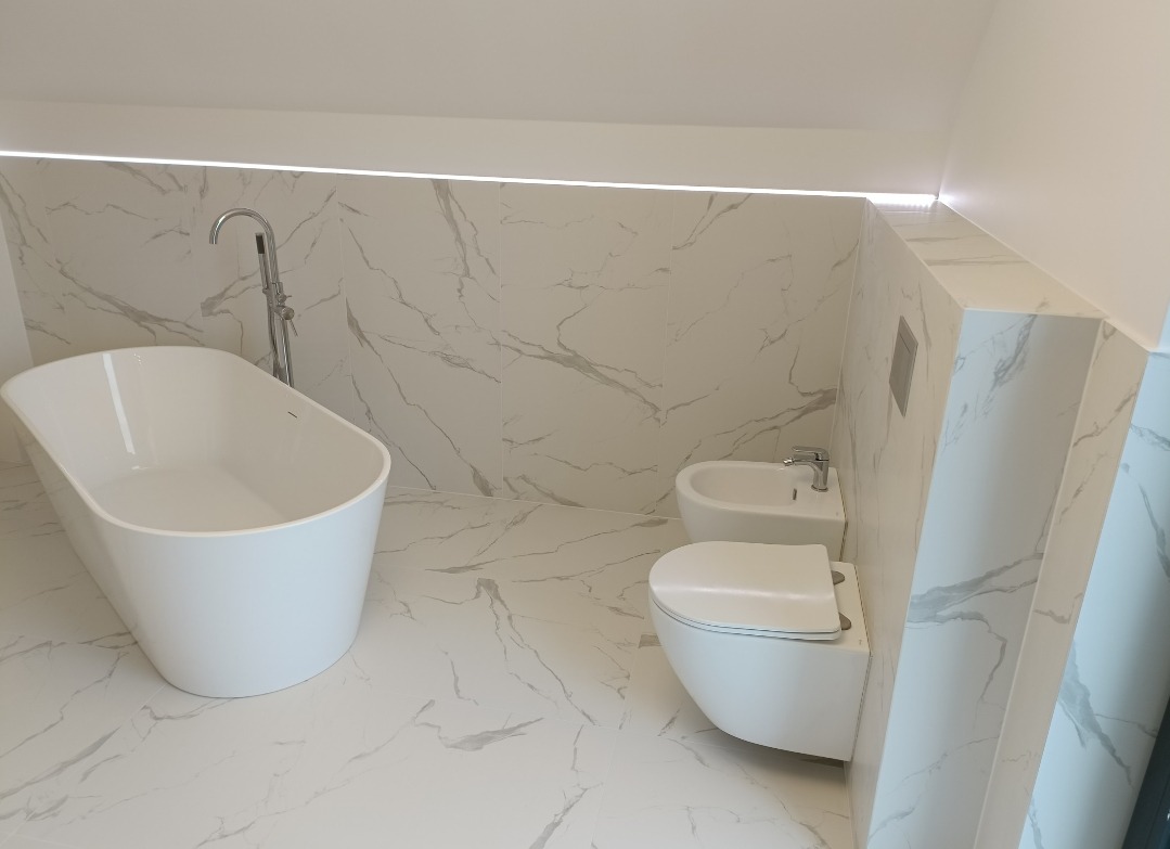 Nowoczesna łazienka w odcieniach bieli z marmurowymi płytkami, wolnostojąca wanna z chromowaną baterią, wisząca toaleta i bidet.