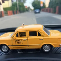 Żółty model taksówki WPT 1313 na desce rozdzielczej samochodu, z widokiem na ulicę w tle.
