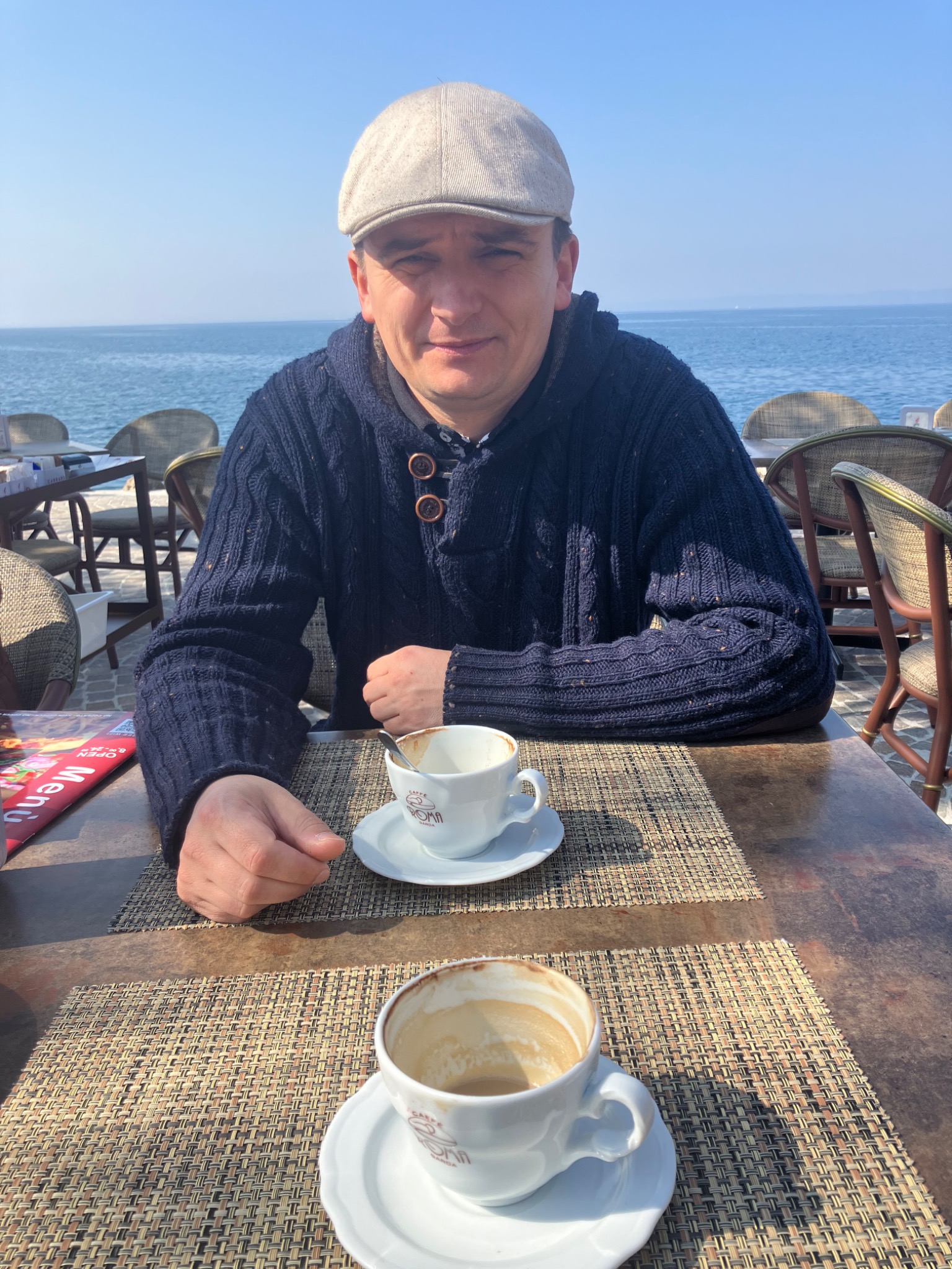 Mężczyzna w czapce siedzi przy stoliku na zewnątrz z widokiem na morze, popijając kawę z filiżanki z logo 'Caffe Roma Garda'.