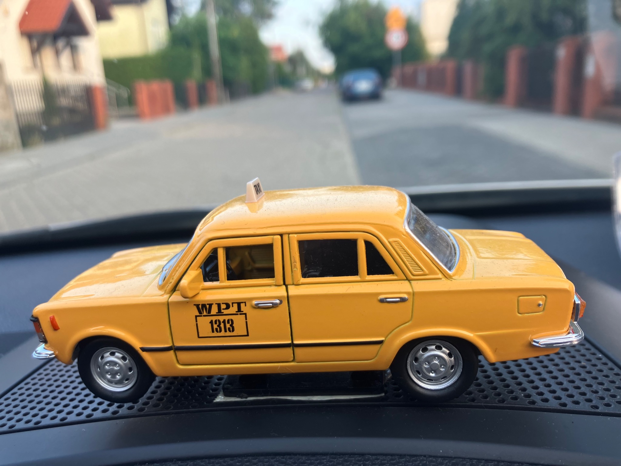 Żółty model taksówki WPT 1313 na desce rozdzielczej samochodu, z widokiem na ulicę w tle.