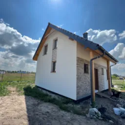 Nowo wybudowany dom jednorodzinny z elewacją częściowo wykończoną białym tynkiem i imitacją cegły, widok z zewnątrz w słoneczny dzień, teren wokół jeszcze niezagospodarowany.