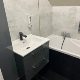 Nowoczesna łazienka z umywalką na szafce w kolorze grafitowym, czarną armaturą, wanną obudowaną czarną mozaiką i szarymi płytkami imitującymi marmur na ścianach, uzupełniona o dozownik mydła.