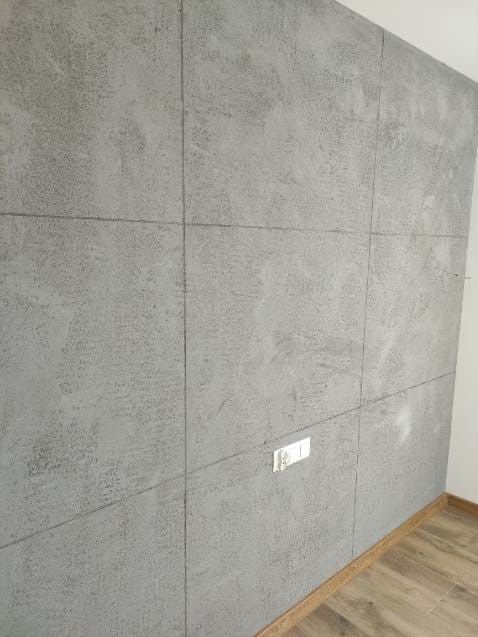 Ściana pokryta szpachlą dekoracyjną imitującą beton, podzielona na kwadratowe panele, z widocznym gniazdkiem elektrycznym i fragmentem podłogi z drewnopodobnych paneli.