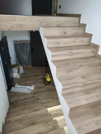 Schody wyłożone panelami imitującymi drewno, z białymi podstopnicami, widoczne narzędzia i materiały budowlane w tle.