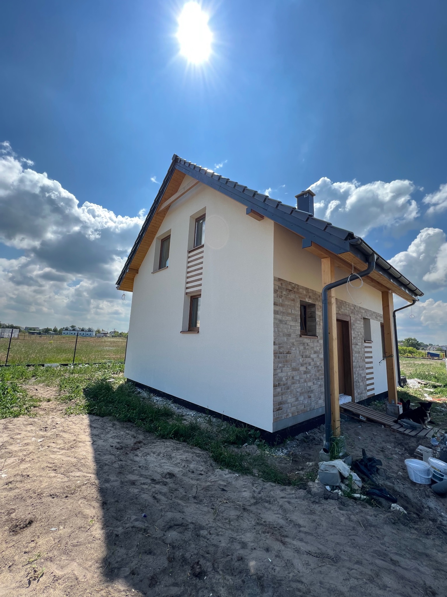 Nowo wybudowany dom jednorodzinny z elewacją częściowo wykończoną białym tynkiem i imitacją cegły, widok z zewnątrz w słoneczny dzień, teren wokół jeszcze niezagospodarowany.
