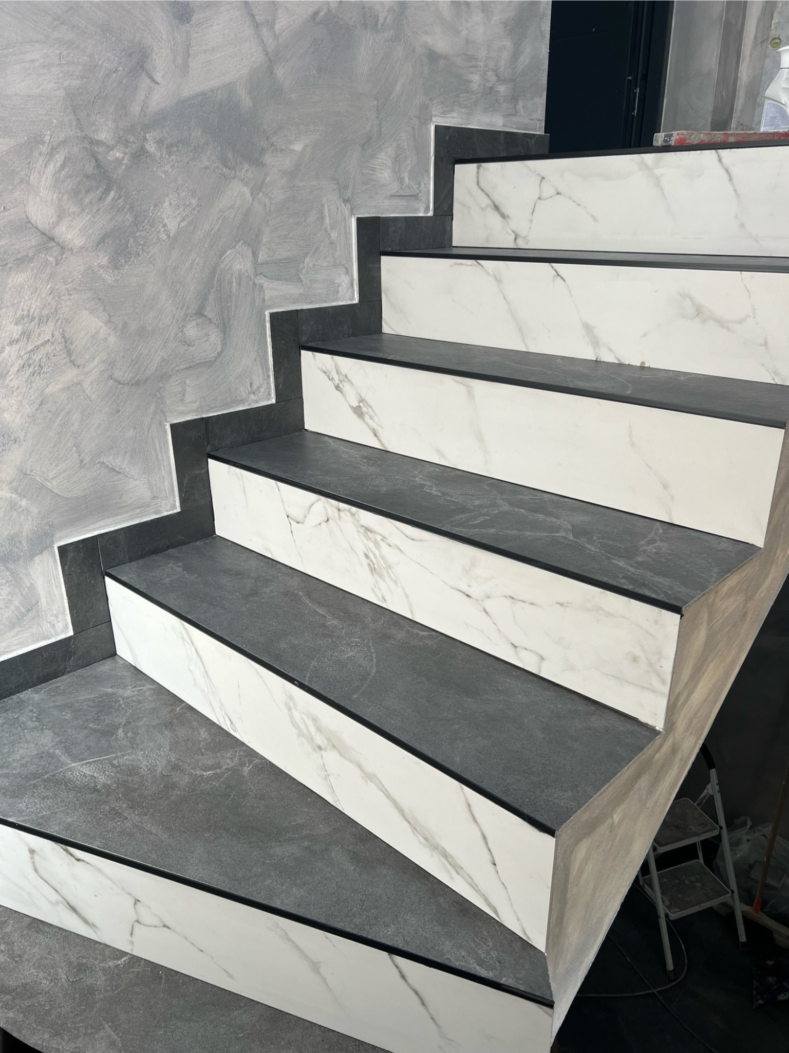 Schody wyłożone szarymi i białymi płytkami imitującymi marmur, z widoczną fakturą ściany i fragmentem drabiny w tle.