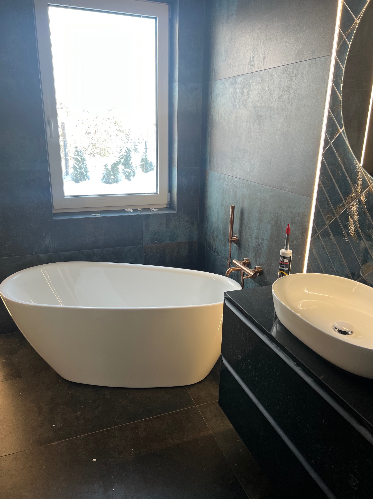 Nowoczesna łazienka z wolnostojącą białą wanną, umywalką nablatową i ciemnymi płytkami, widoczny aplikator silikonu na blacie umywalki.