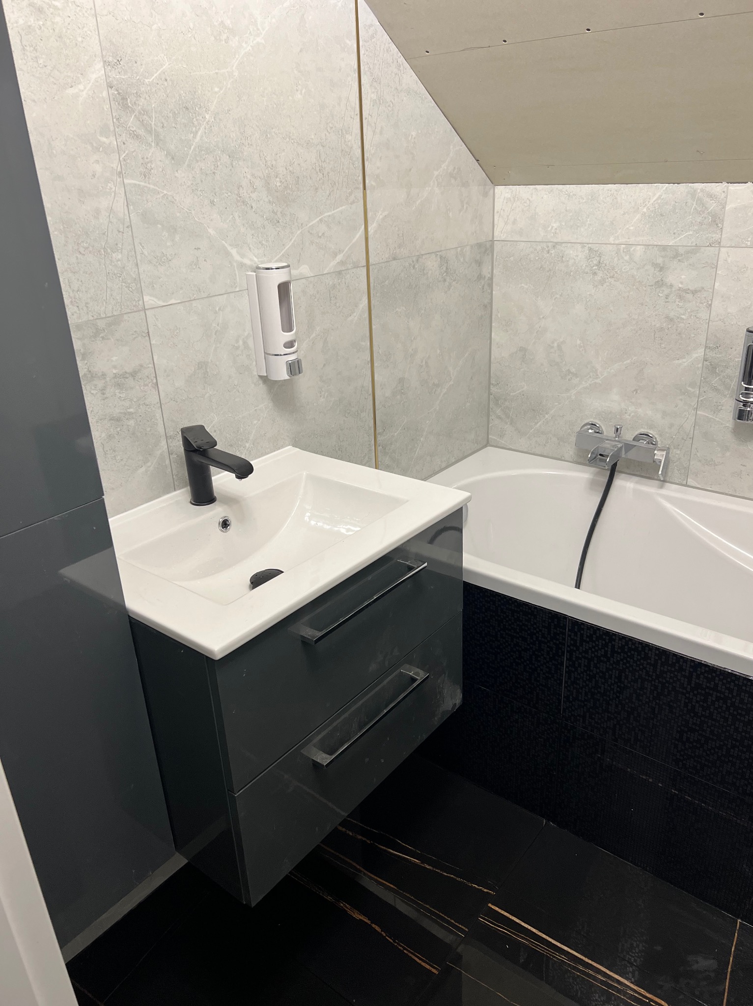 Nowoczesna łazienka z umywalką na szafce w kolorze grafitowym, czarną armaturą, wanną obudowaną czarną mozaiką i szarymi płytkami imitującymi marmur na ścianach, uzupełniona o dozownik mydła.