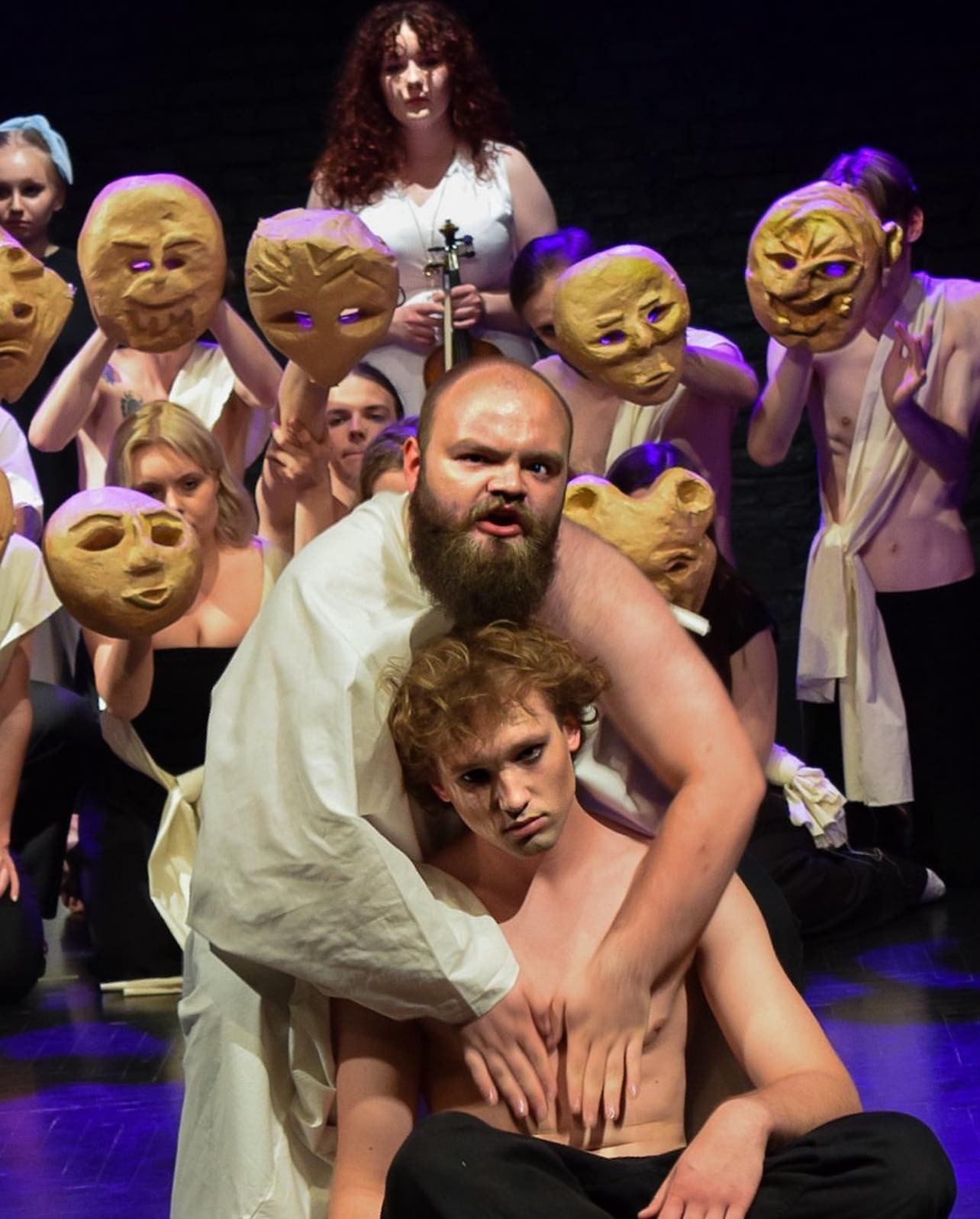 Scena teatralna z aktorami w kostiumach i maskach, centralnie mężczyzna z brodą obejmuje młodego aktora bez koszuli, w tle pozostali aktorzy trzymają maski.