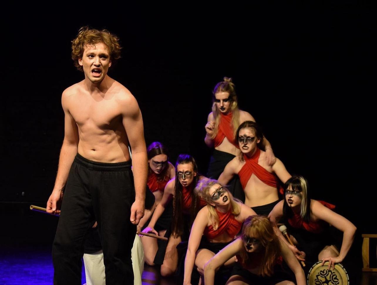 Scena teatralna: grupa aktorów w kostiumach i makijażu plemiennym, z bębnem i pałeczkami, w trakcie przedstawienia.